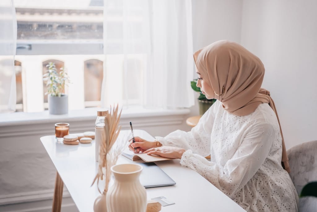 A woman in a hijab writing at a sun-lit desk near a window in a cozy room.